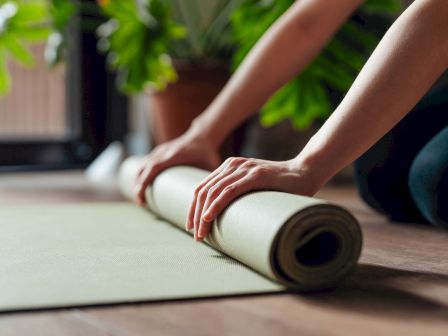 Hands rolling up a yoga mat on a wooden floor, with green plants in the background.