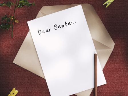 A letter to Santa, surrounded by a pencil, coffee cup, gingerbread cookie, and festive decorations on a red surface.