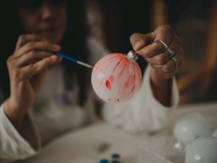 A person is painting a white ornament with red paint. They are using a small brush to create designs on the surface.