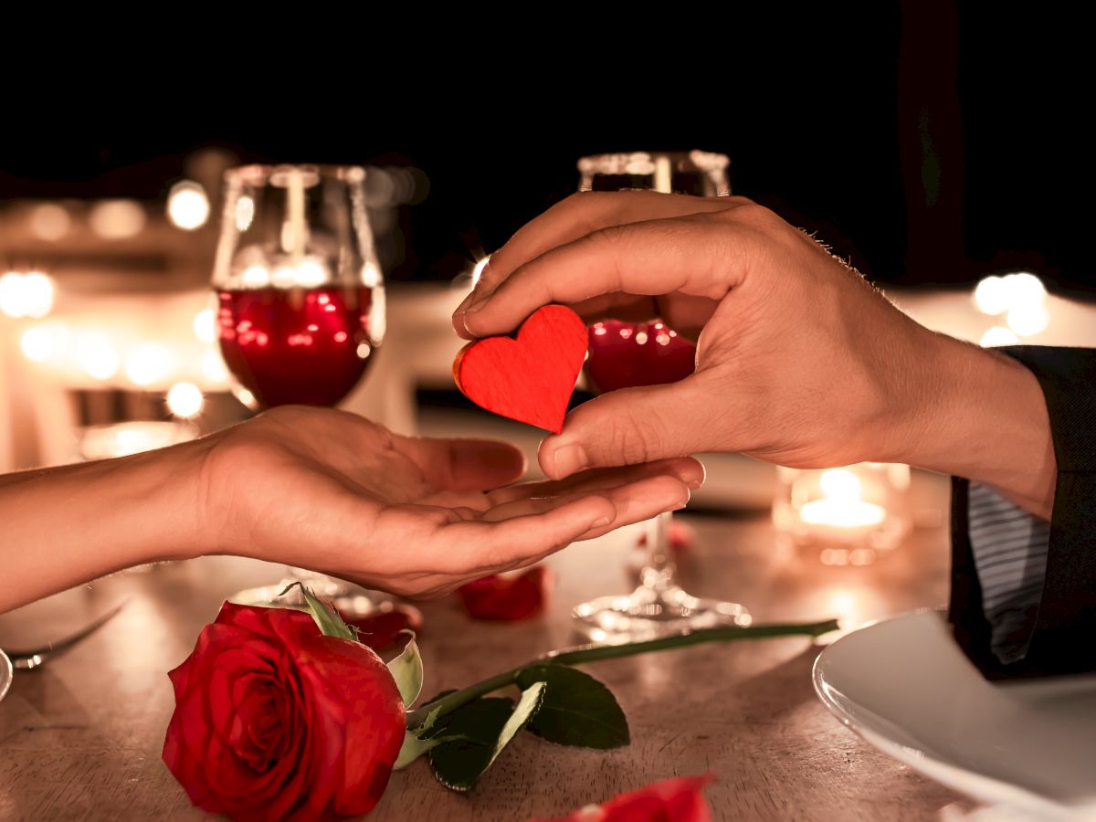 A romantic setting with red wine, a rose, and hands exchanging a small red heart, surrounded by a cozy candlelit ambiance.