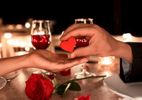 A romantic dinner setting with two wine glasses, a red rose, and a hand offering a small red heart to another hand.