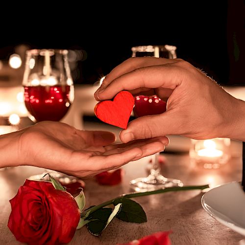 A romantic dinner setting with two wine glasses, a red rose, and a hand offering a small red heart to another hand.