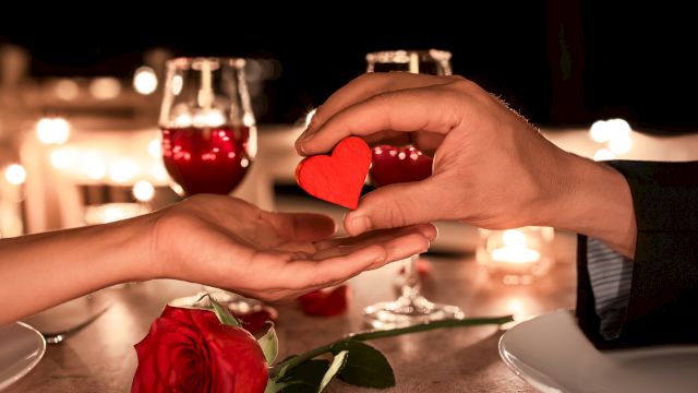 A romantic dinner setting with two wine glasses, a red rose, and a hand offering a small red heart to another hand.