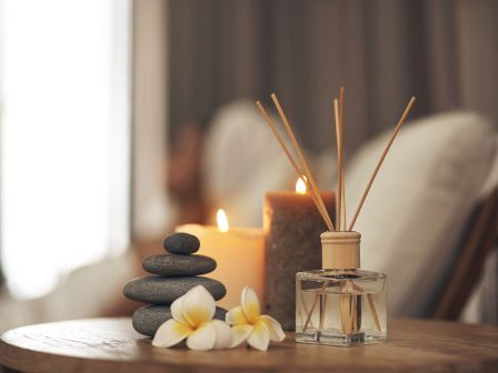 The image shows a spa scene with stacked stones, candles, frangipani flowers, and a diffuser on a wooden surface, creating a calming atmosphere.