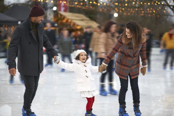 A family ice skating, holding hands, and smiling outdoors with festive lights in the background.