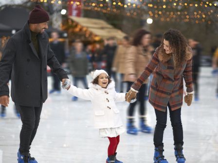 A family ice skating together, holding hands, with festive lights and a crowd in the background, all wearing warm winter clothes.