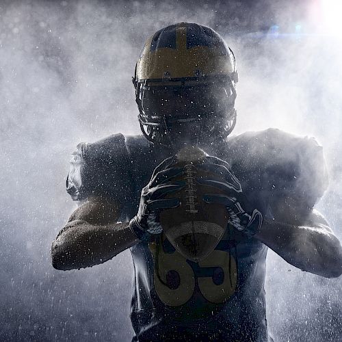 The image shows a football player wearing a helmet, holding a football, with dramatic lighting and mist surrounding them.