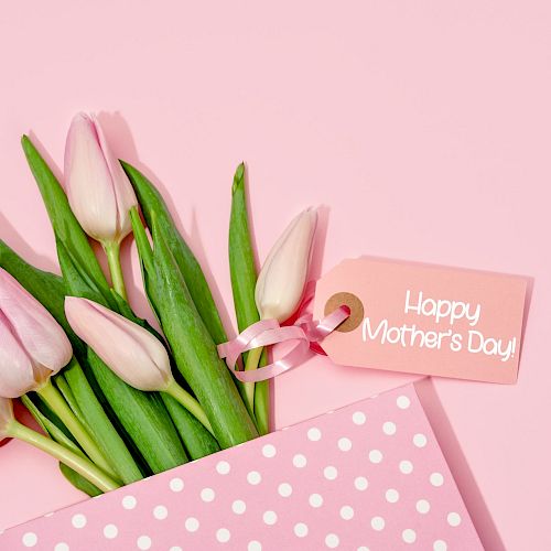 Pink tulips in a polka dot envelope with a "Happy Mother's Day" tag on a pink background.