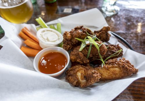A plate of fried chicken wings with sliced green onions, accompanied by carrot sticks, celery, ranch, and hot sauce, served with a drink.
