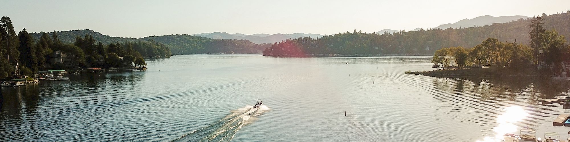 A boat moves across a calm lake surrounded by trees and distant hills under a clear sky with the sun shining.