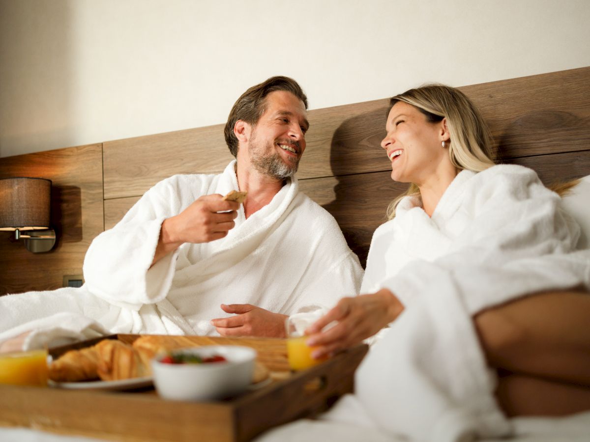 A couple in bathrobes enjoys breakfast in bed, sharing pastries and juice, and smiling at each other in a cozy setting.