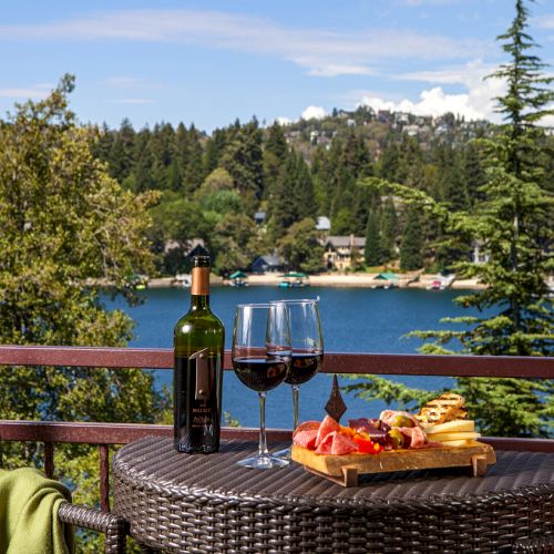 A table with wine, glasses, and a charcuterie board on a balcony overlooking a scenic lake with trees and hills in the background.