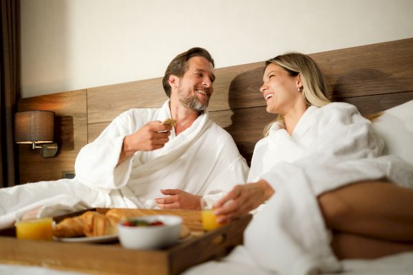 A couple in bathrobes enjoying breakfast in bed with croissants, juice, and fruit, sharing a lighthearted moment in a cozy setting.