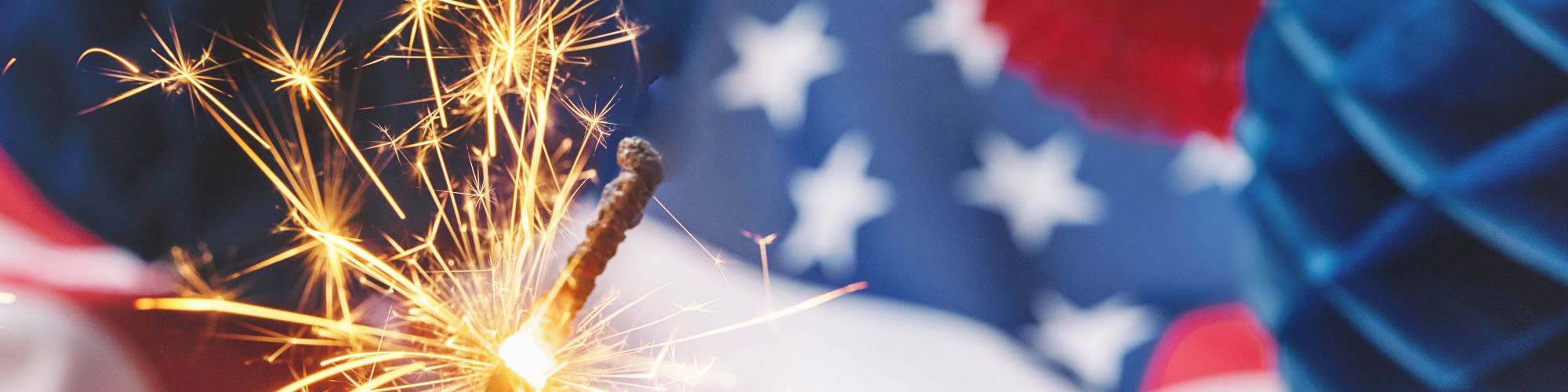 A lit sparkler is in front of a blurred US flag and colorful decorations, creating a festive atmosphere.