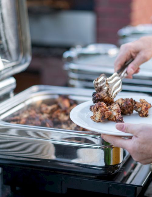 A person with tongs is placing food onto a plate from a buffet station, next to a stack of white plates.