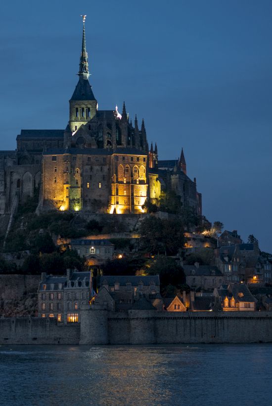 The image shows Mont Saint-Michel, an illuminated island commune in France, featuring a medieval abbey and surrounded by water at dusk.
