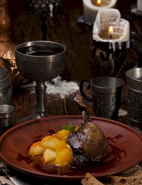 A rustic setting with a metal goblet, pewter cups, candles, and a plate of cooked pears with sauce, evoking a medieval feast.