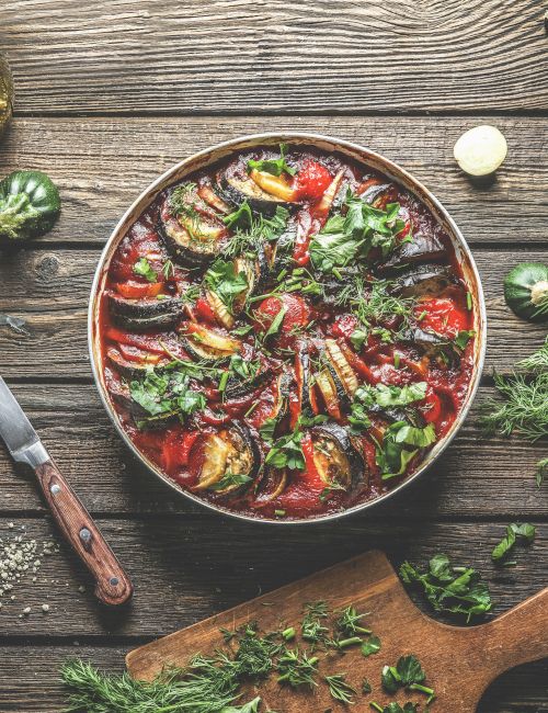 A dish with sliced vegetables like eggplant, zucchini, and tomatoes, garnished with herbs, on a wooden table with a knife and herbs nearby.