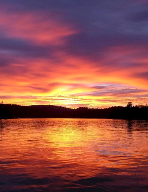 A vibrant sunset over a calm lake, with silhouettes of trees and hills under a colorful sky reflecting on the water's surface.