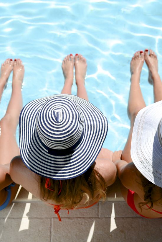 Three people wearing sun hats sit at the edge of a pool, with their legs dipped in the water.