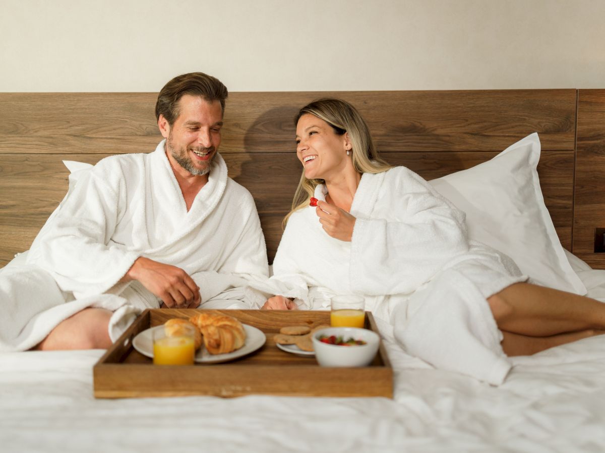 A couple in bathrobes enjoys breakfast in bed with orange juice, croissants, and fruit, sharing smiles and a relaxed moment together.