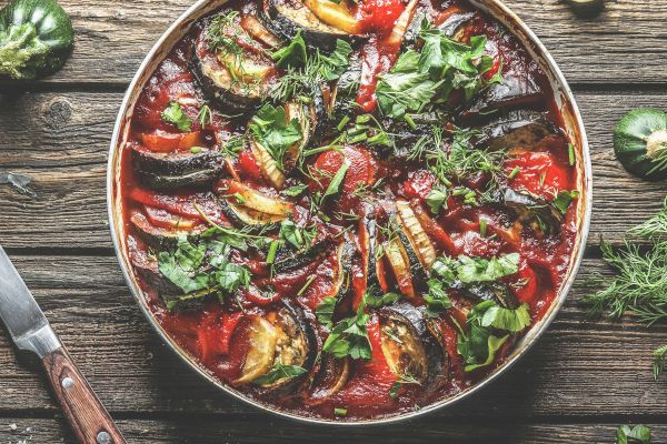 A colorful dish with sliced vegetables, possibly ratatouille, garnished with herbs on a wooden table, surrounded by a knife and more herbs.